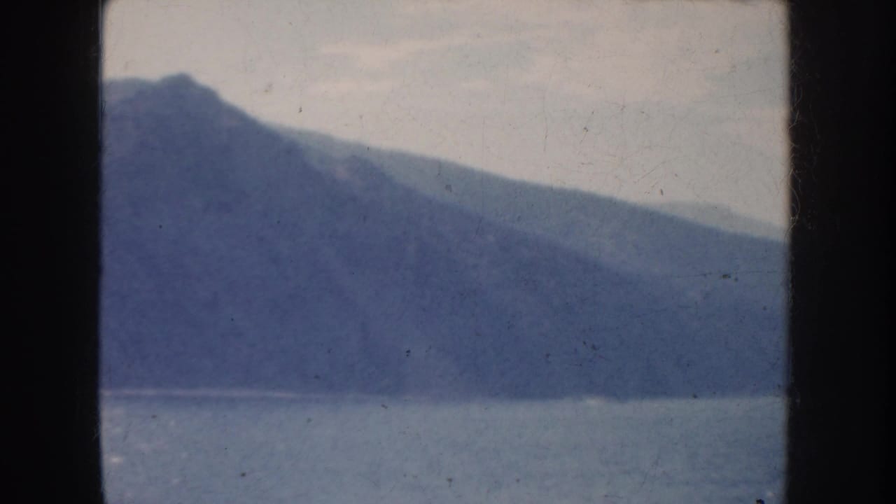 1952 California home-movie view of a hazy mountain shoreline across calm blue water, framed by dark window edges.