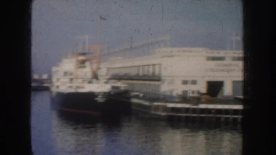 1952 California 8mm harbor view showing a dark-hulled vessel moored near a pier and large waterfront building.
