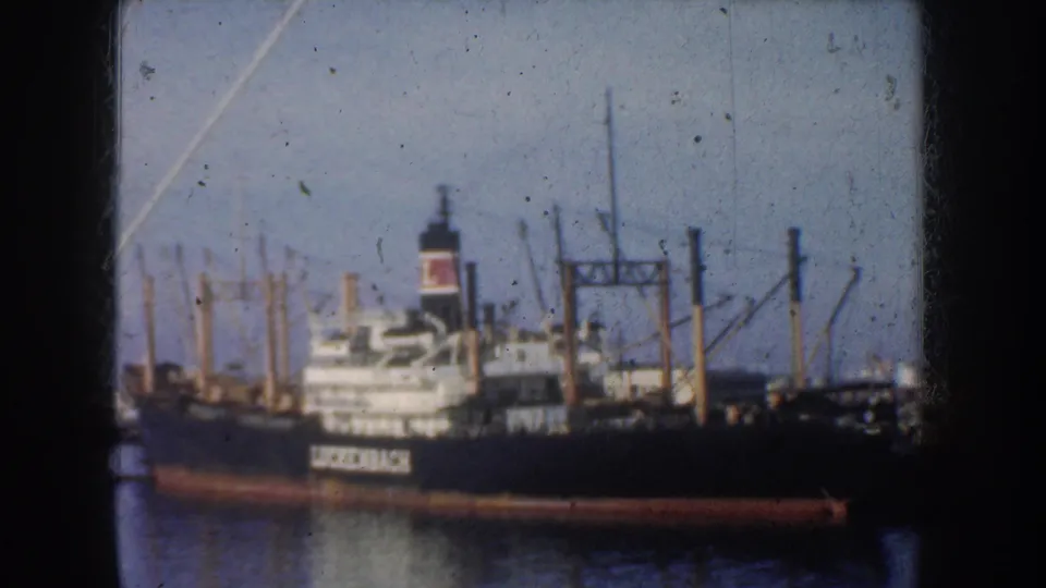 1952 California 8mm frame of the Luckenbach cargo ship at dock, with harbor cranes, masts and calm water visible.