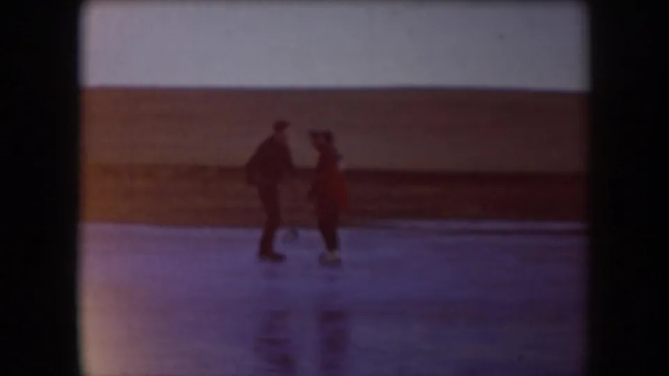 Two adults skate across a frozen North Dakota lake, one in dark clothing and one in a red coat, in soft 1950s home-movie footage.