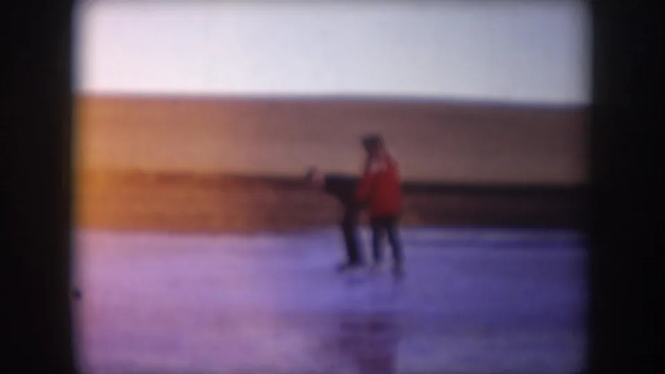 A skater in a bright red coat follows a darker-clothed skater across the frozen lake in 1953 North Dakota home-movie footage.