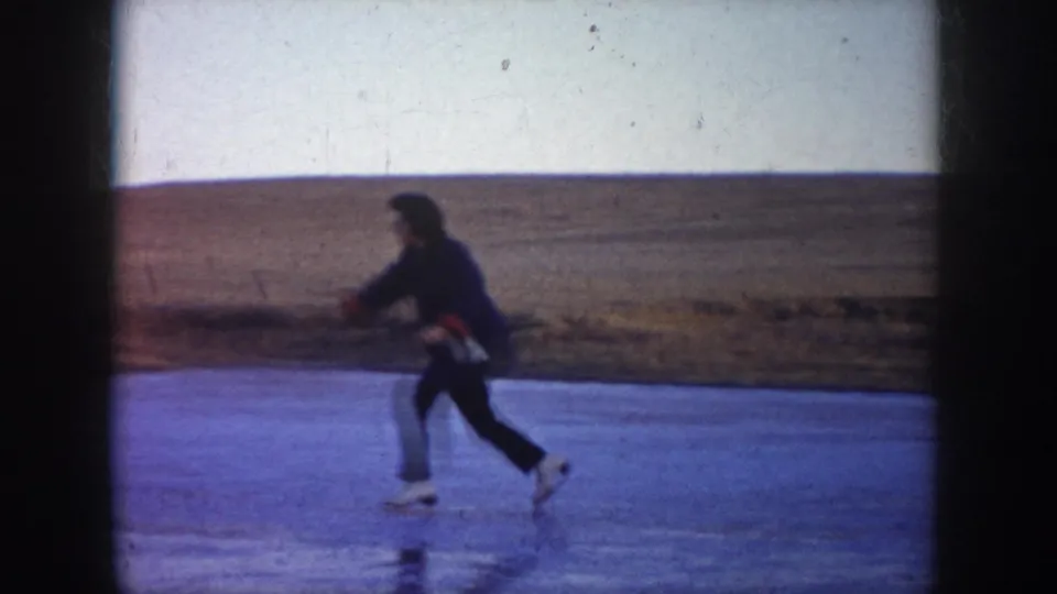 A person in winter clothing skates across the frozen lake, filmed in a soft 8mm home-movie frame in North Dakota.
