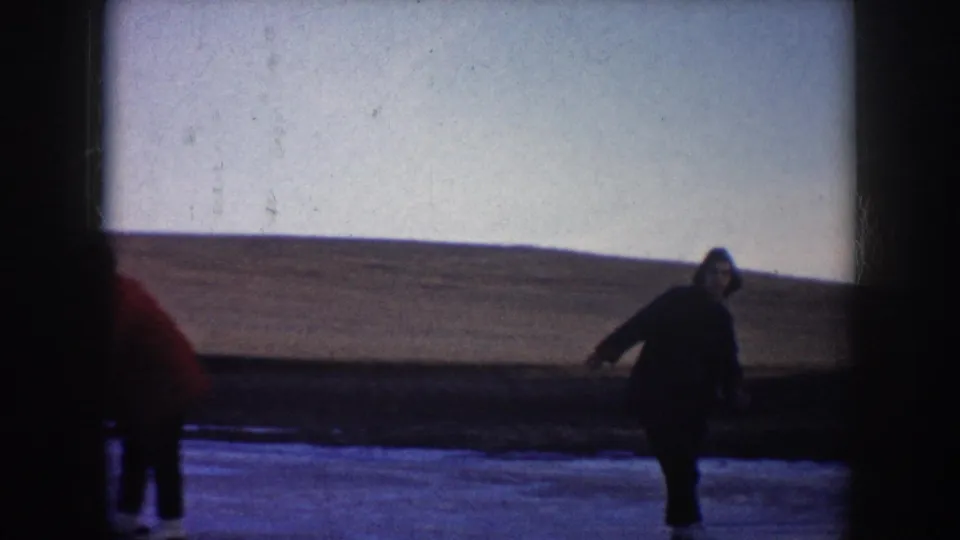 A skater in a dark coat glides across the frozen lake, framed by dark 8mm gate edges and a wide rural horizon.