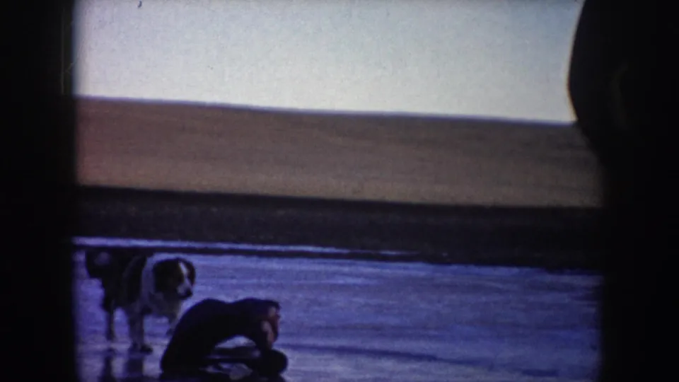 Home-movie frame showing a dog on frozen water and a person crouched low on the ice in rural North Dakota.