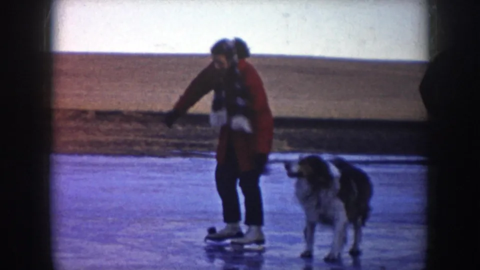 1953 North Dakota 8mm frame of a skater in a red coat on a frozen lake with a large dog alongside.