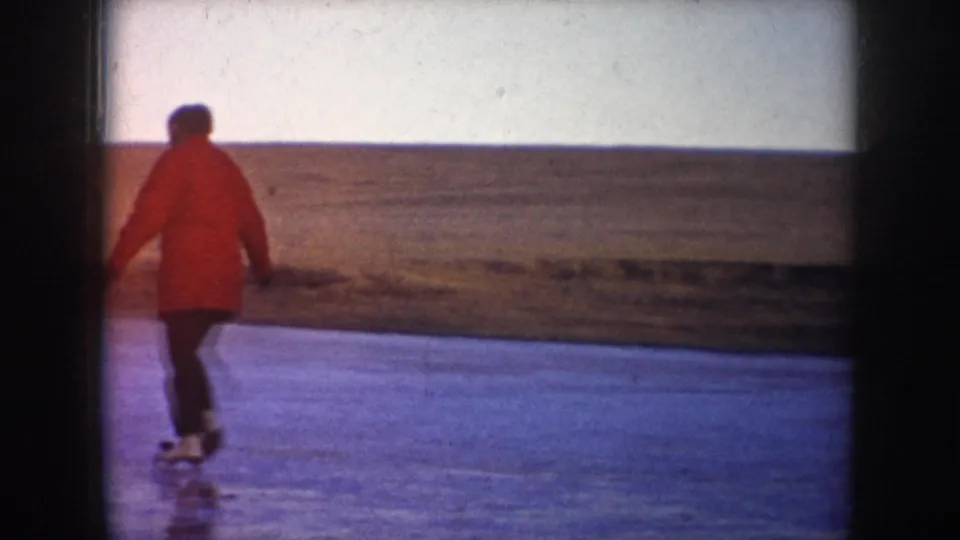 A red-coated skater moves away across open ice in 1953 North Dakota, with a flat rural horizon and pale sky behind them.