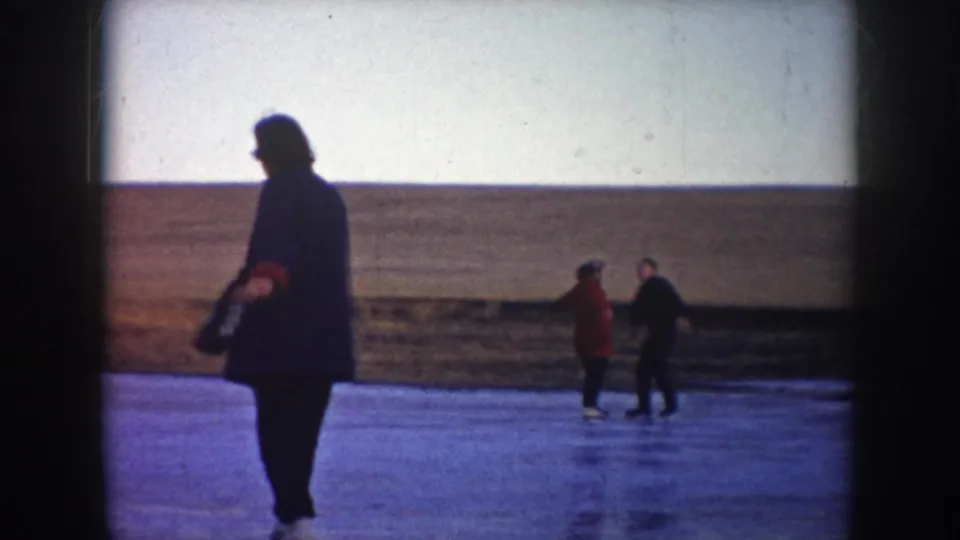 A dark-clothed adult stands near the frozen surface as two distant skaters, including one in red, meet on the ice in North Dakota home-movie footage.