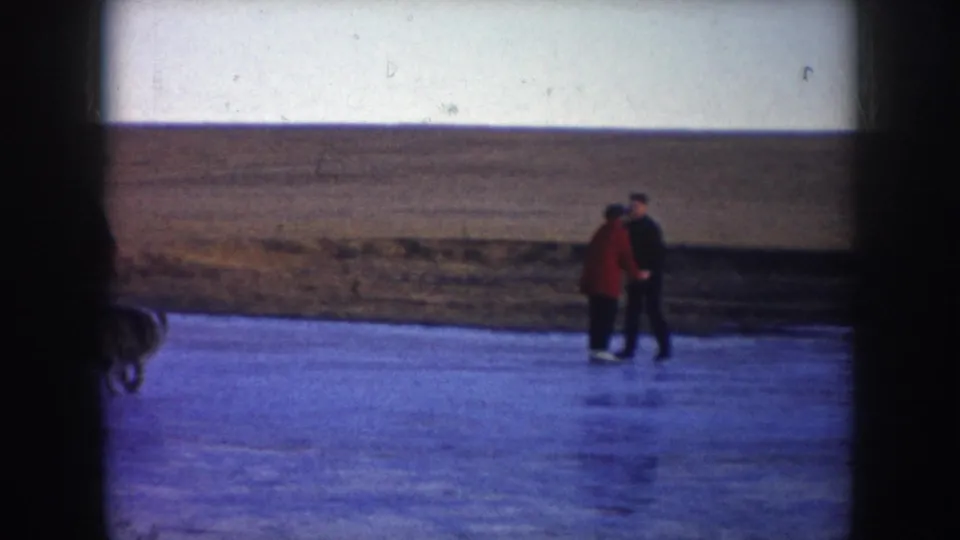 Two adults, one in a red coat and one in dark clothing, skate or stand together on outdoor ice as a dog passes partly through the frame.