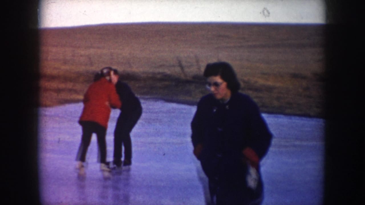 1953 North Dakota home-movie frame showing two adults skating on a frozen pond with a third adult in dark clothing nearby.