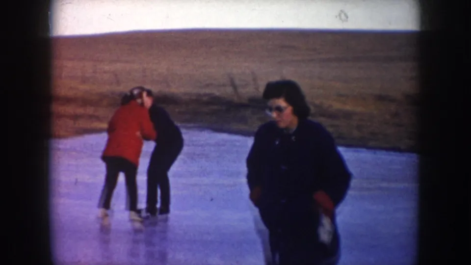 1953 North Dakota home-movie frame showing two adults skating on a frozen pond with a third adult in dark clothing nearby.