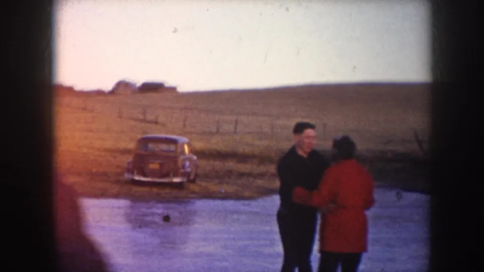 The skating couple pauses on the frozen pond, with a period car parked on the rural bank behind them.