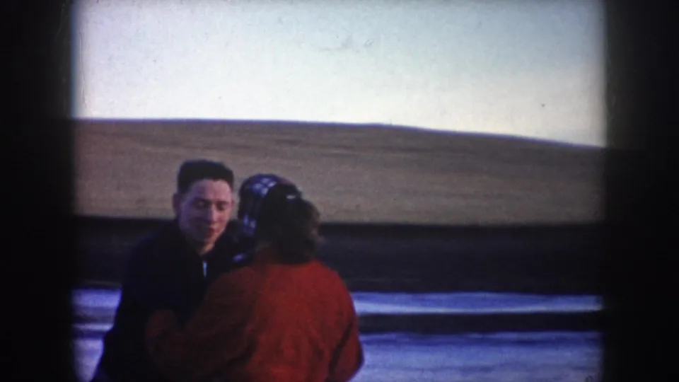 Close home-movie view of the skating couple on the frozen pond, with the man facing camera and the woman in a red coat turned away.