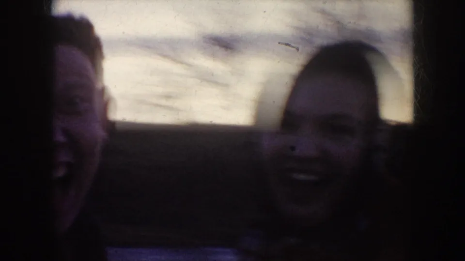 Two adults laugh near the camera in a dark, grainy 1950s North Dakota winter home-movie frame.