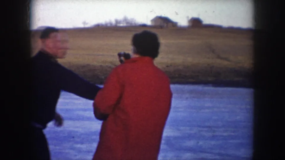 A man and a woman in a red coat stand on a frozen lake in rural North Dakota, with fields and distant buildings beyond.