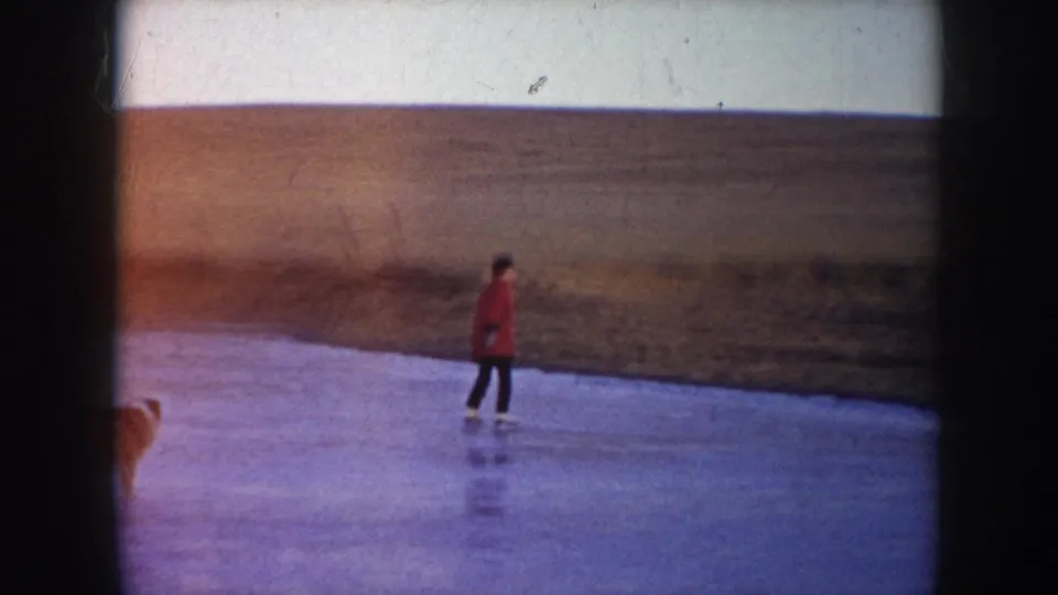 1953 North Dakota frame of a skater in a red coat gliding over a frozen lake with rural fields behind.