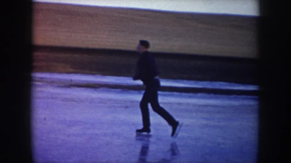 1953 North Dakota home-movie frame showing a dark-clothed skater on a frozen lake near rural fields.
