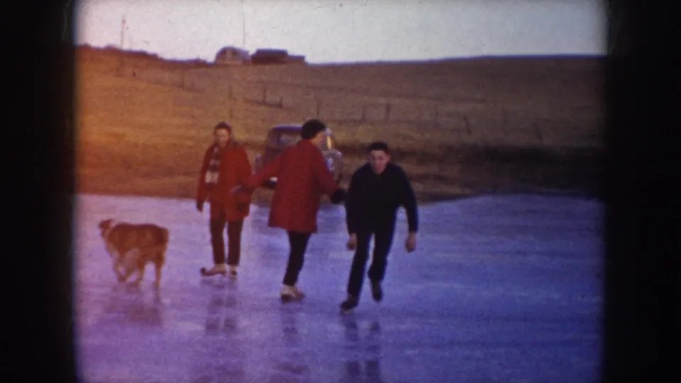 1953 North Dakota frame showing multiple skaters and a dog on a frozen lake, with a parked car near the shore.
