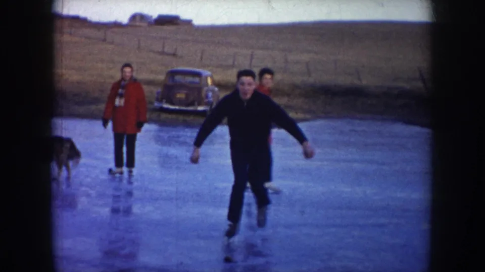 1953 North Dakota home-movie frame showing adults skating on a frozen pond, with a vintage car on the bank and open rural fields beyond.