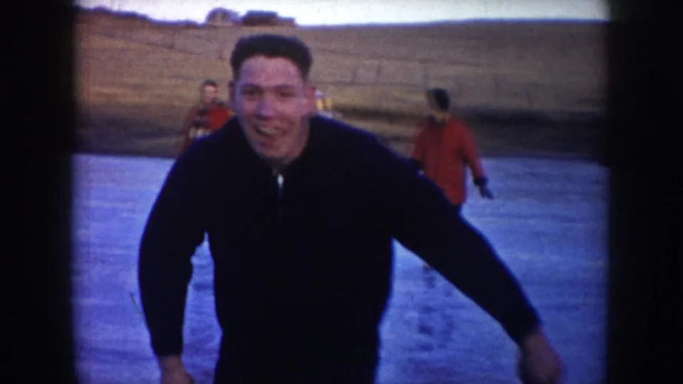 A smiling adult man skates toward the camera in this 1953 North Dakota winter home-movie frame, with other skaters behind him.
