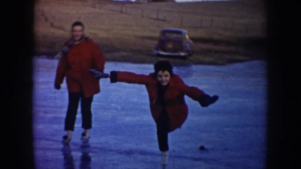 Two adults in red winter coats skate on a frozen pond in North Dakota, with one extending arms for balance near a parked car in the background.