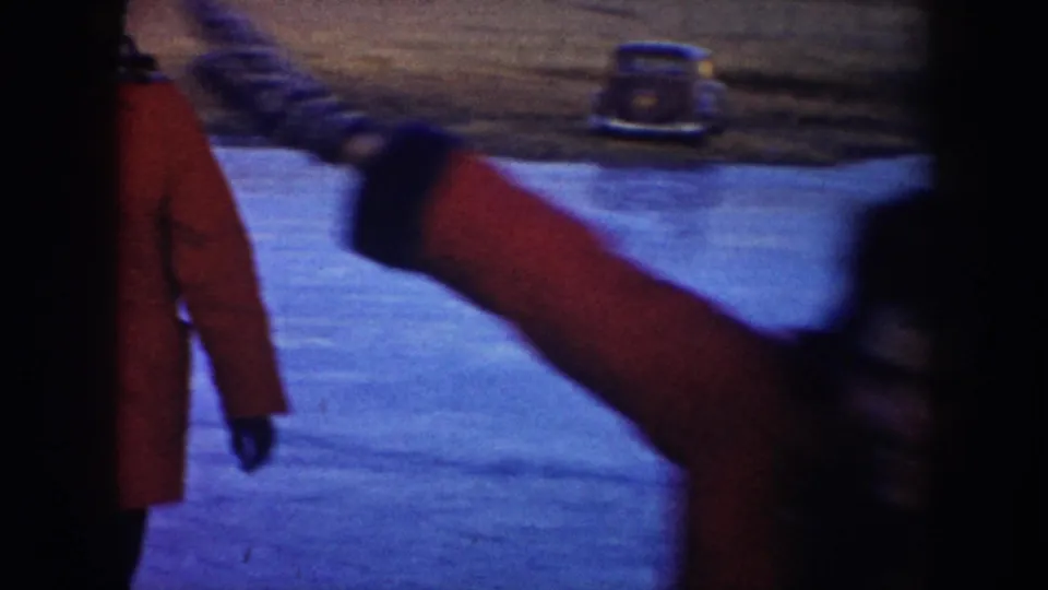 A close, blurred home-movie frame catches a red-coated skater passing the camera, with the frozen pond and parked car still visible behind.