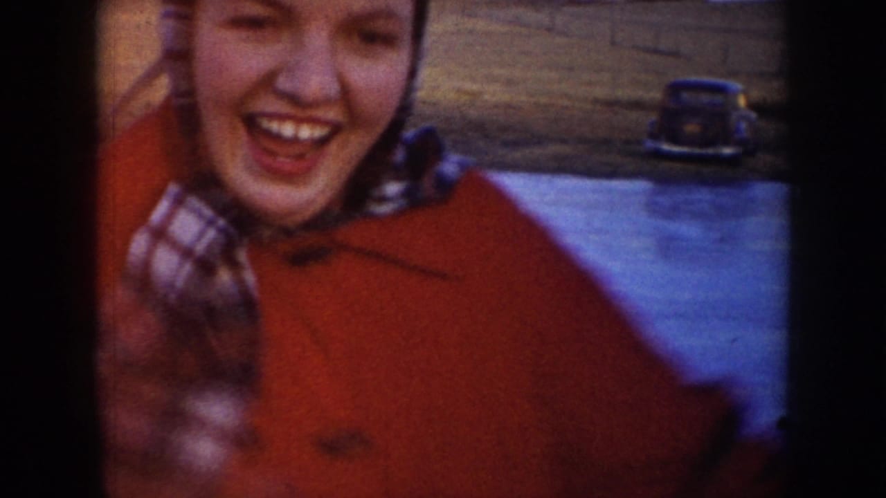 In supported 1953 North Dakota home-movie footage, a woman in a red coat and plaid scarf smiles near the camera on the ice.