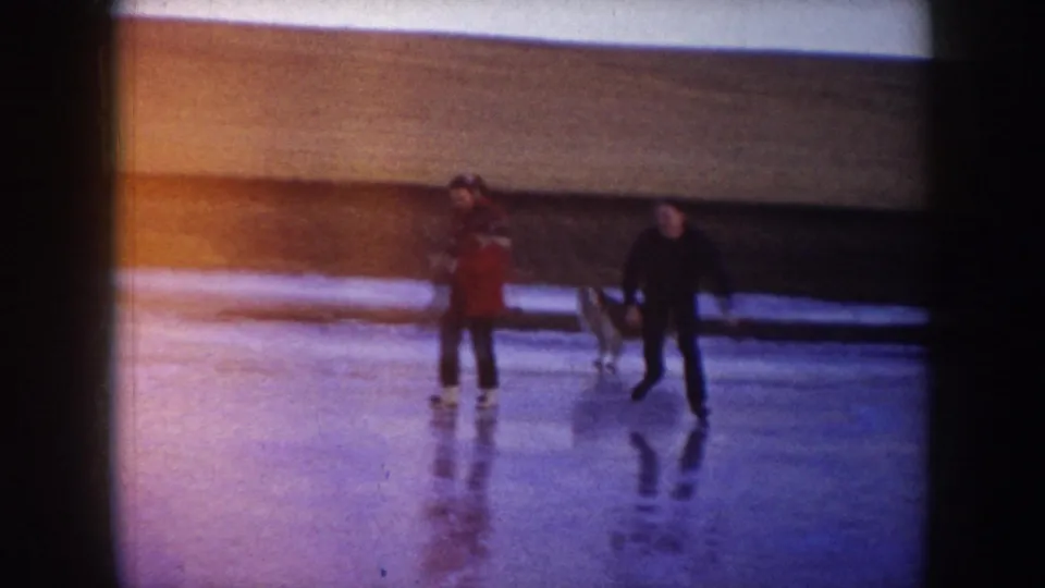 Two skaters and a dog move across smooth ice in the North Dakota winter landscape, with reflections visible on the lake surface.