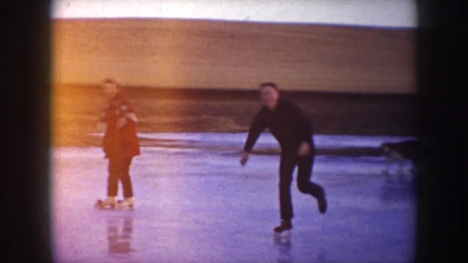 A woman in red pauses on the ice as a man skates past, with a dog partly visible near the edge of the 8mm frame.