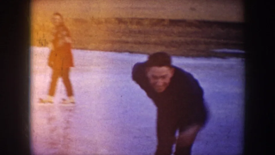 A man in dark clothing bends toward the camera on skates while the woman in red remains behind him on the frozen lake.