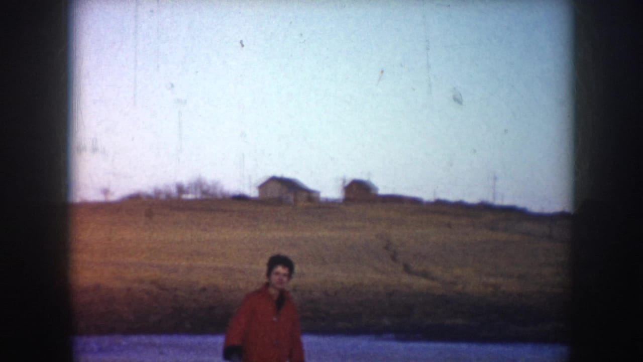 1953 North Dakota home-movie frame showing a skater in a red coat on a frozen pond with rural fields and distant houses.