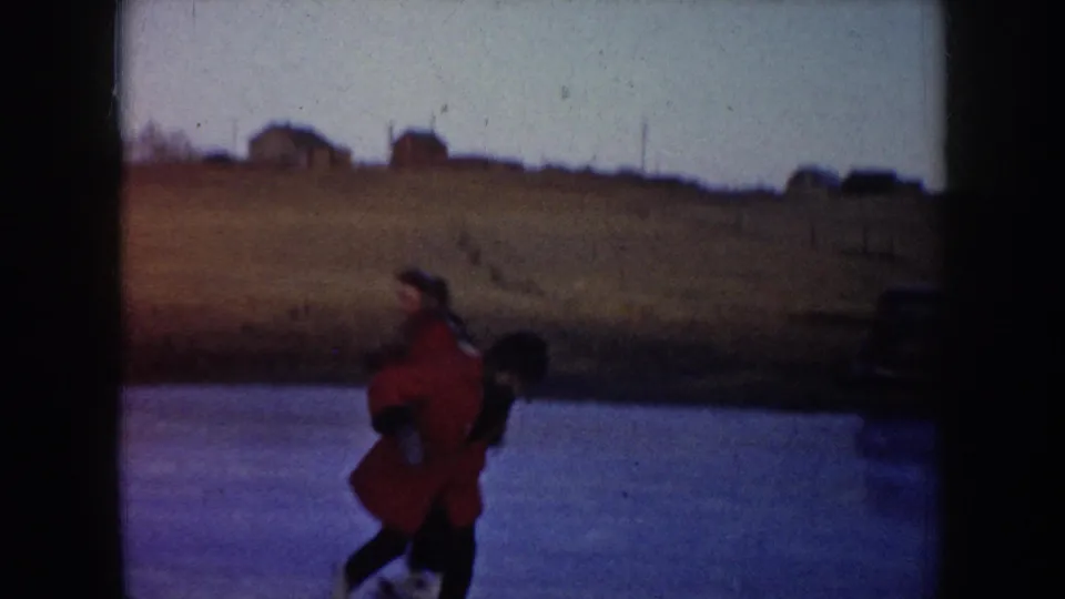 Two skaters move together on a frozen pond in rural North Dakota, with fields and distant houses visible in the background.