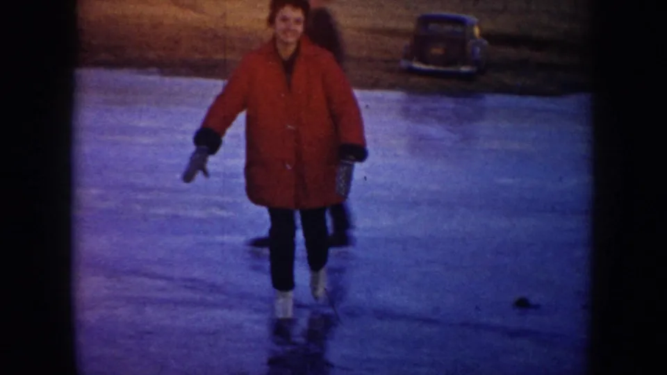 A skater in a bright red coat smiles while skating toward the camera on a frozen pond, with a parked car behind.