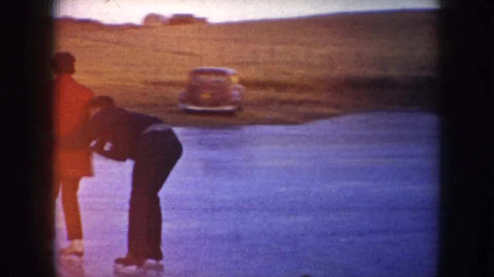 Two skaters pause on the frozen pond in rural North Dakota, with a mid-century car parked near the field behind them.