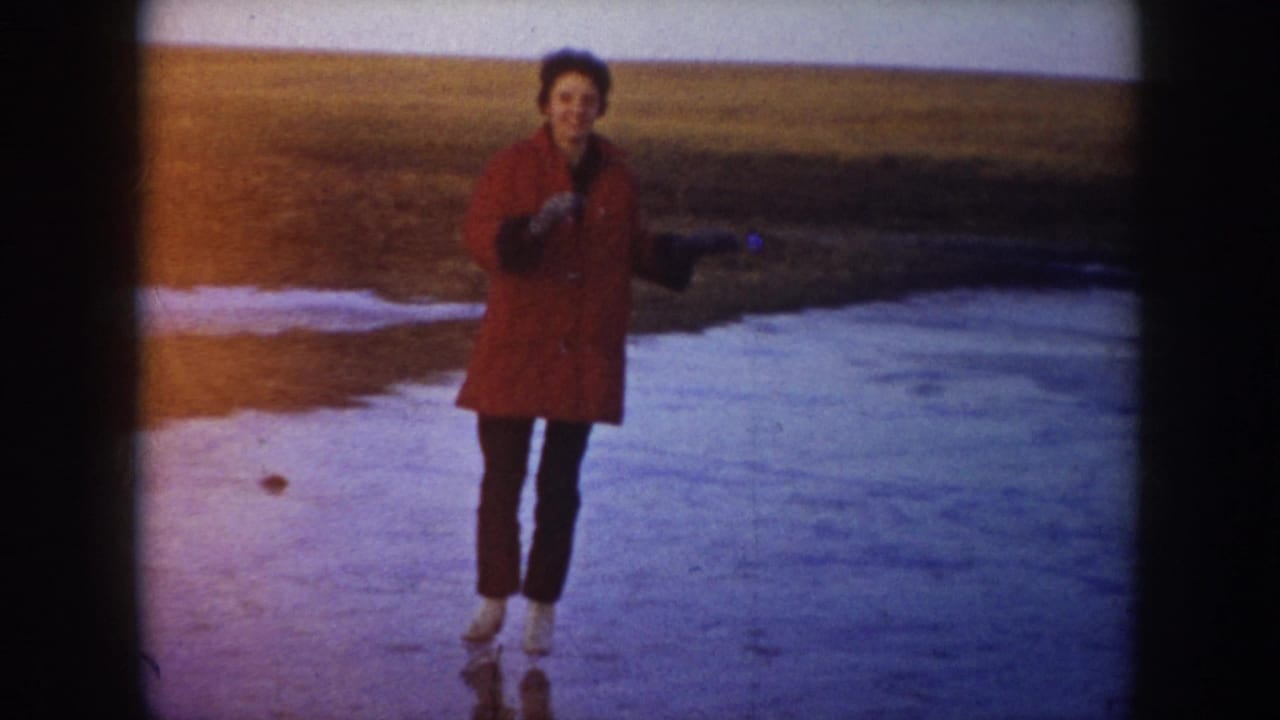 1953 North Dakota home-movie frame of a skater in a red coat balancing on a frozen lake near open fields.