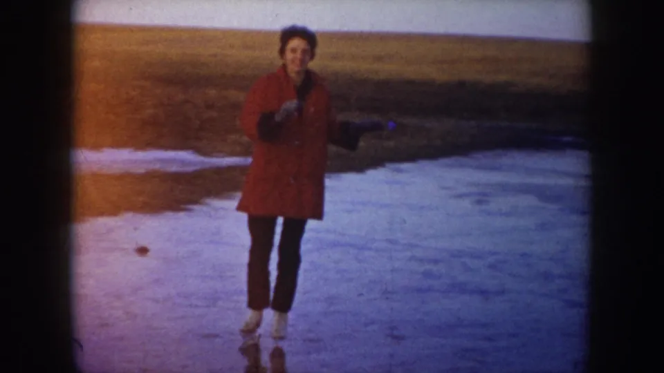 1953 North Dakota home-movie frame of a skater in a red coat balancing on a frozen lake near open fields.