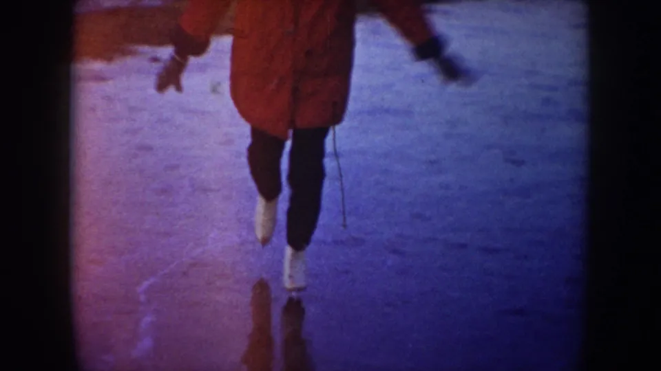 Close 8mm frame of the red-coated skater's legs and white skates gliding over blue frozen lake ice.