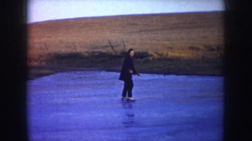Wide 1953 North Dakota home-movie frame showing a distant skater in a dark coat on a frozen lake beneath open fields.