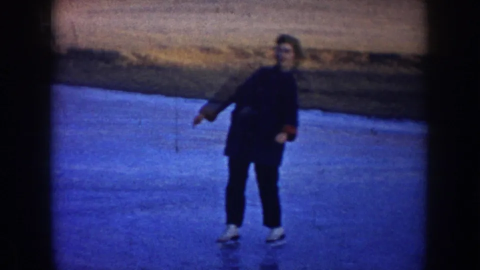 1953 North Dakota 8mm frame of a skater in a dark coat balancing on blue lake ice near a brown shoreline.