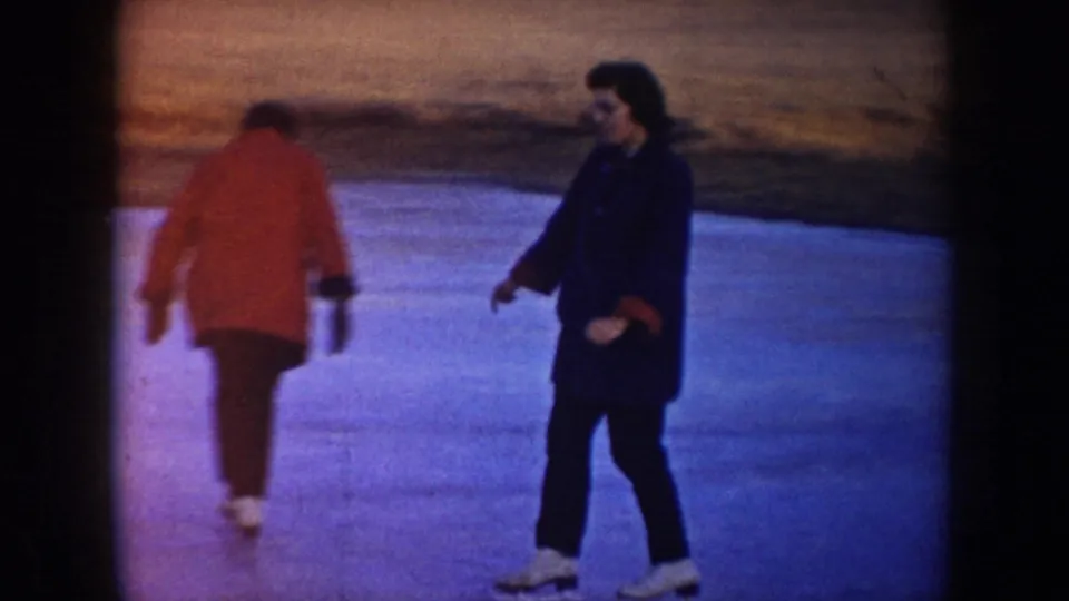 1953 North Dakota 8mm frame showing two skaters on blue ice, one in a red coat and one in a dark coat, with fields behind them.