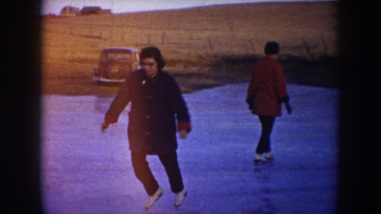1953 North Dakota home-movie frame with a dark-coated skater in front, a red-coated skater at right, and a parked car near the field edge.