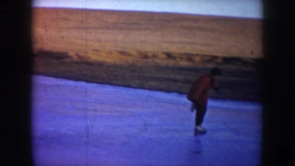 1953 North Dakota 8mm frame showing a red-coated skater leaning forward on blue ice, with a grassy bank and open field beyond.