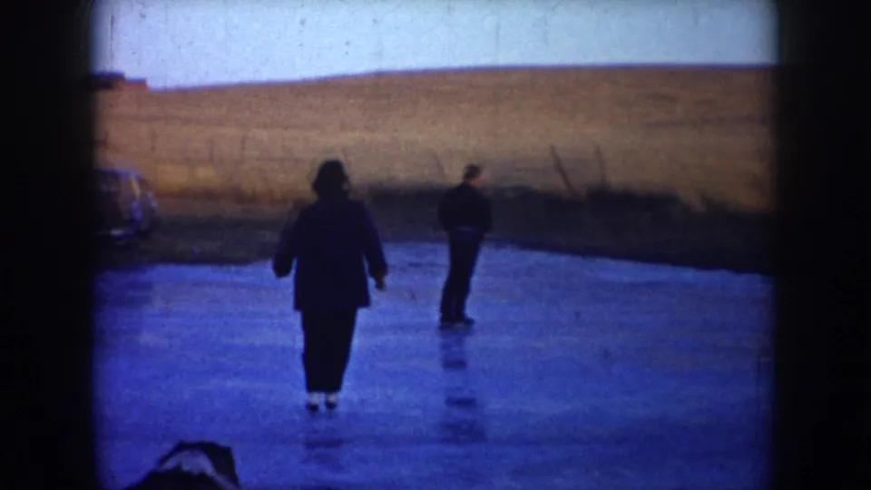 1953 North Dakota home-movie frame with two skaters seen from behind on a frozen pond, open fields, and part of a parked car at left.