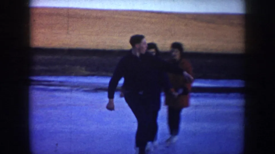 Three skaters move close together on the ice, with one red coat standing out against the rural winter background.