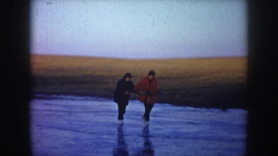 Two people, one in a red coat and one in a dark coat, hold hands while skating on a rural North Dakota pond.