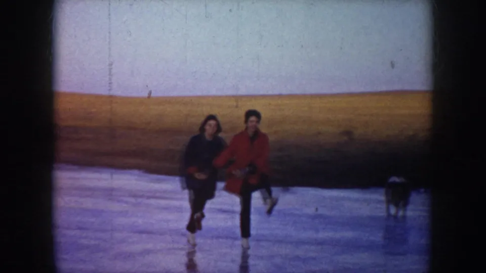 Two adults in winter coats glide on a frozen North Dakota lake, with open fields behind them and a small dark form at the right edge.