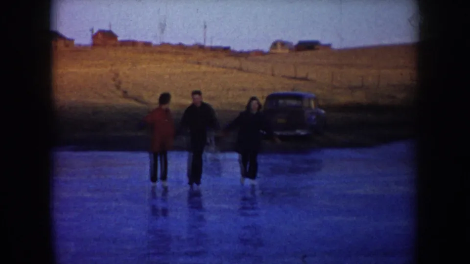 Three adults walk or slide on a frozen lake in North Dakota, with a parked mid-century car visible behind them near open fields.