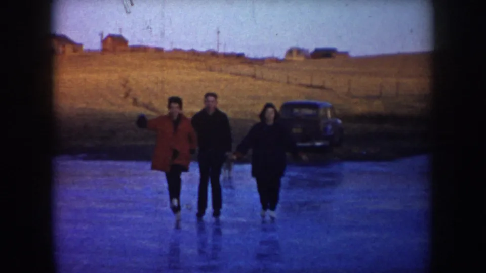 Three adults on a frozen North Dakota lake face the camera, one in a red coat gesturing, with a parked car and rural field behind them.