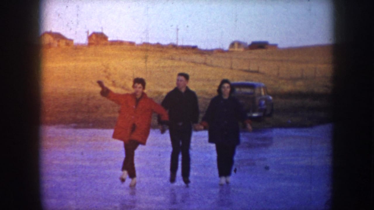 In this 1953 North Dakota 8mm frame, three young adults skate hand in hand across a frozen rural pond, with one wearing a red coat and a rounded-body car parked near distant houses.