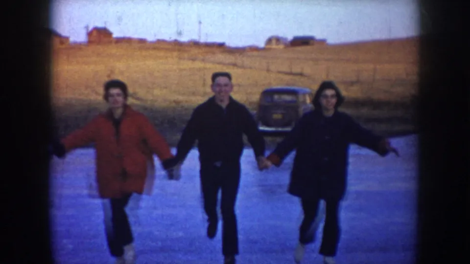 Three young adults skate toward the camera on a frozen pond in North Dakota, with a red coat, dark jackets, a parked car, and open rural land visible behind them.