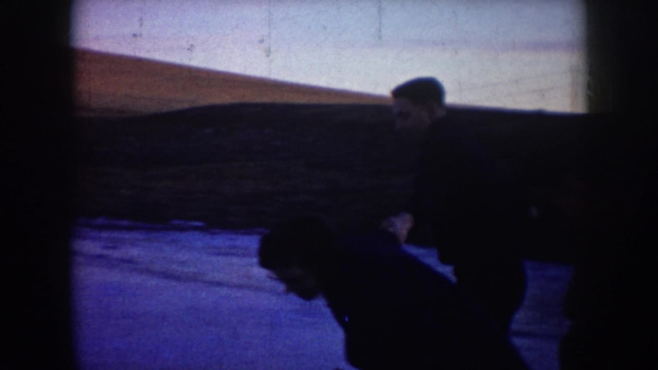 1953 North Dakota home-movie frame showing two adults in dark winter clothing skating on a frozen pond.